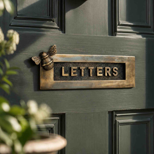 Unlacquered aged brass bumble bee "LETTERS" letter plate fitted to a dark sage green front door, featuring raised lettering and a decorative bee motif.