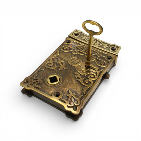 Angled view of unlacquered aged brass floral rim lock with key inserted, showing engraved floral pattern.