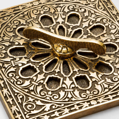Close-up angled view of the aged brass square air vent showing the ornate openwork design, textured background and decorative border detailing.
