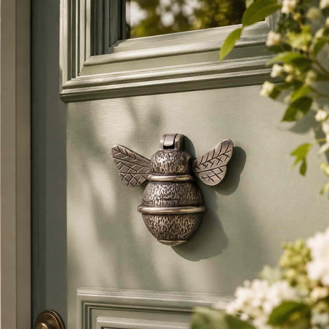 Aged brass bumble bee door knocker fitted to a sage green front door surrounded by garden foliage