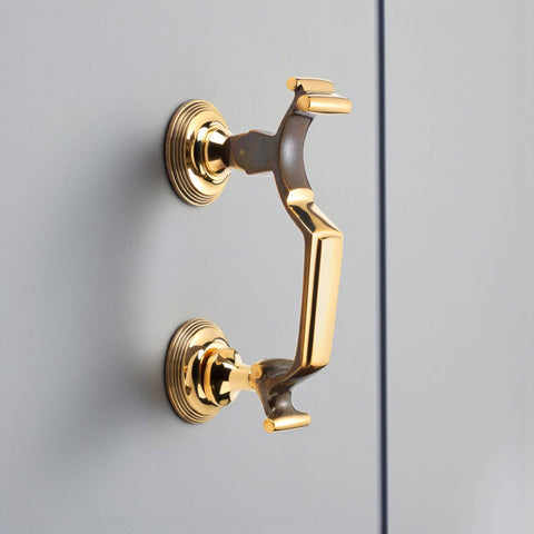 Unlacquered aged brass reeded doctor door knocker mounted on grey door, angled side view showing circular backplates and solid brass detail.