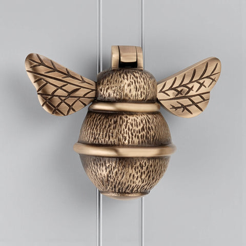 Solid brass bumble bee door knocker in antique brass finish, front view showing aged detailing and textured body on grey door.