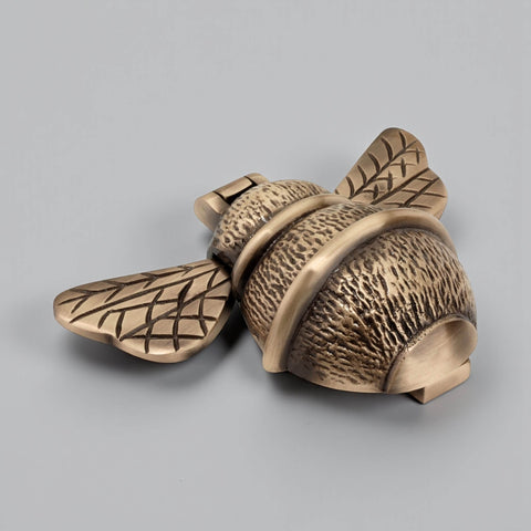 Antique brass bumble bee door knocker shown at an angle, highlighting textured body, smooth bands and engraved wing detail.