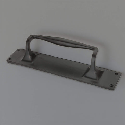 Angled view of black cast iron door pull handle on rectangular backplate with four countersunk fixing holes.
