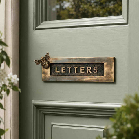 Unlacquered aged brass bumble bee "LETTERS" letter plate installed on a sage green front door, showcasing its naturally aged living brass finish and raised lettering.