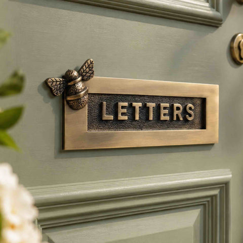 Close-up of an antique brass bumble bee "LETTERS" letter plate installed on a sage green front door, showcasing the textured background and raised lettering.