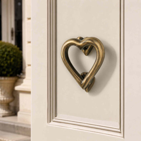 Solid brass antique brass heart door knocker mounted on a cream front door, showcasing its traditional aged finish and elegant heart-shaped design.