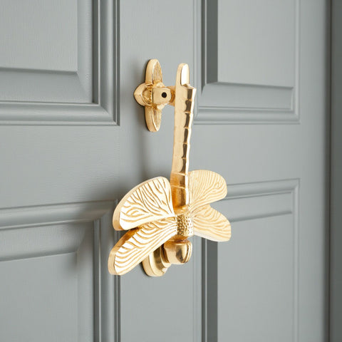 Side view of polished brass dragonfly door knocker on a grey door showing detailed textured wings.