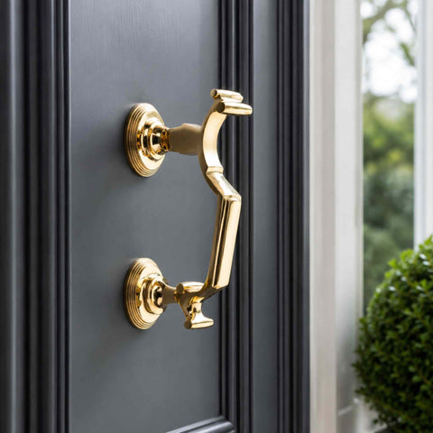 Angled close-up of a polished brass reeded doctor door knocker mounted on a dark grey front door, showing the substantial projection, reeded roses and solid brass construction.
