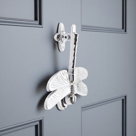Side view of polished chrome dragonfly door knocker on a grey door showing detailed textured wings.