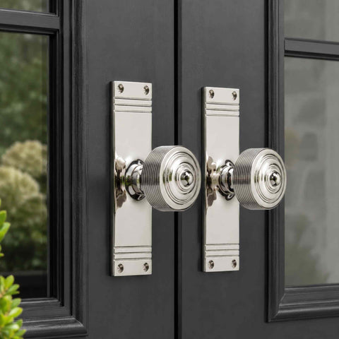 Pair of polished nickel reeded door knobs mounted on matching tall backplates fitted to black double doors, creating a classic and symmetrical entrance feature.