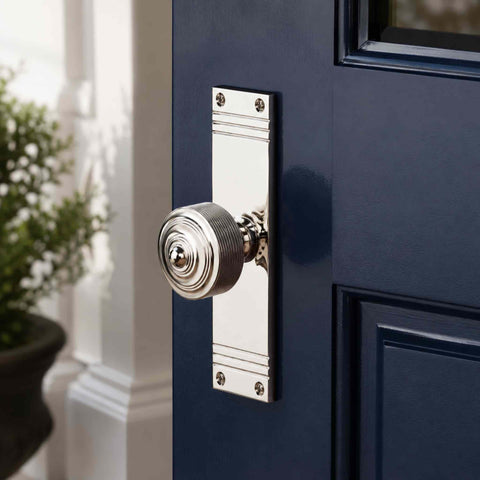 Polished nickel reeded door knob on tall backplate fitted to a navy blue panelled door, demonstrating the contrast between the bright finish and dark door.