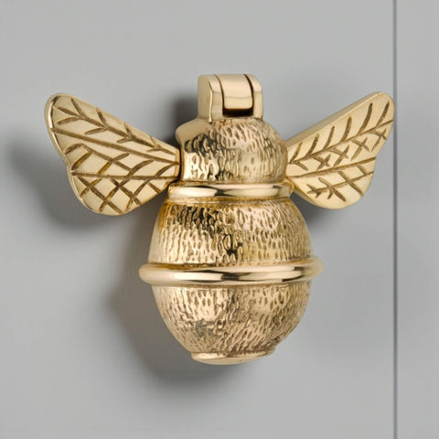 Solid brass bumble bee door knocker in unlacquered polished brass, front view showing detailed wings and textured body on grey door.