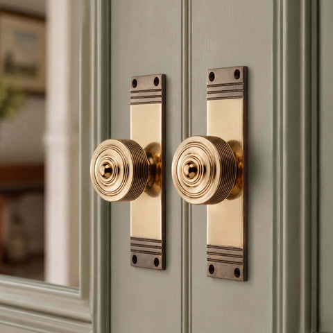 Reeded aged brass door knob mounted on a tall backplate fitted to a sage green panelled door, highlighting the characterful aged finish and period-inspired styling.