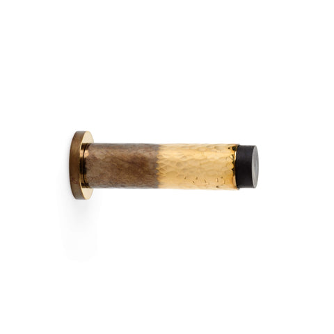 Solid brass hammered cylindrical door stop in aged brass and polished brass finishes, featuring a round mounting plate and black rubber tip, side view on white background.