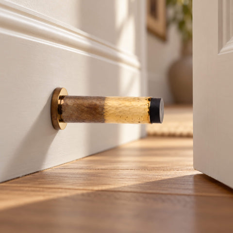 Solid brass hammered cylindrical door stop with contrasting aged and polished brass finishes, installed on a white skirting board beside an open door in an elegant hallway with wooden flooring.