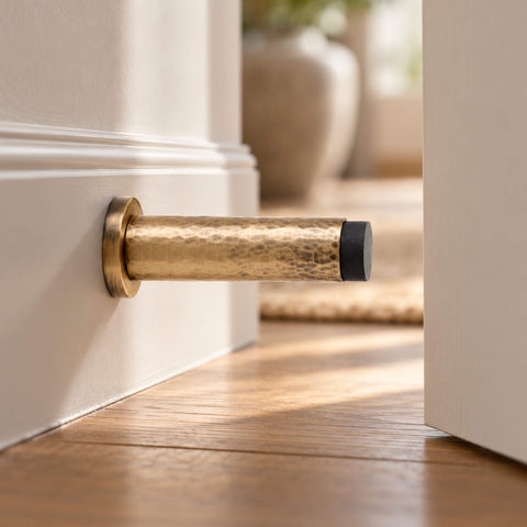 Solid brass hammered cylindrical door stop in antique brass finish, mounted on a white skirting board beside an open door in an elegant hallway with wooden flooring.