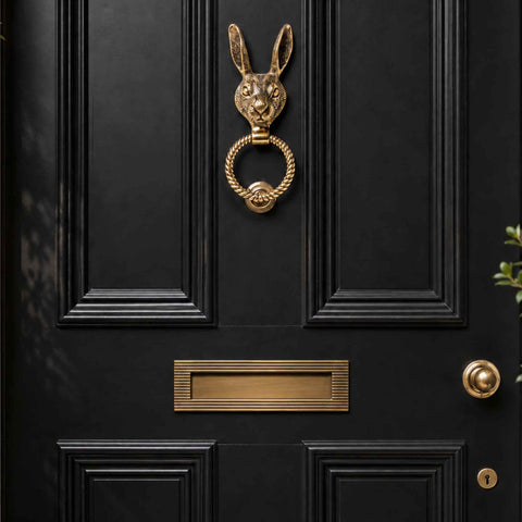 Aged brass hare door knocker with reeded ring installed on a black front door above a matching letter plate